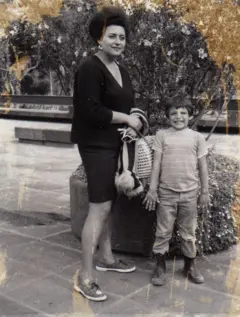 Alejandra Jáidar lleva puestos una falda y un saco negros y unos zapatos planos. A su lado, sonríe su hijo, que lleva unas botas, una camisa de rayas y un pantalón. Atrás de ellos hay un árbol pequeño