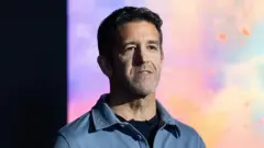 約翰・特納斯身穿藍色襯衫、內搭黑色 T 恤，站在一面由藍、紫、黃、橙與粉紅色交織而成的繽紛背景前