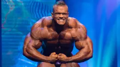 Dallas McCarver no palco do Arnold Classic em um fundo azul, ostentando braços musculosos.