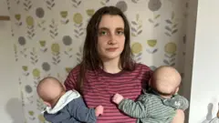 Ella Marcham tem cabelos castanho-escuros e olha para a câmera enquanto segura dois bebês; ela usa uma camiseta listrada vermelha, e os bebês vestem azul-marinho e verde