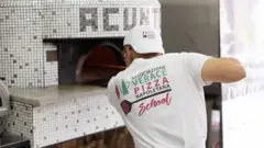 Uma pessoa colocando uma pizza em um forno. Ela usa uma camisa com o nome da AVPN