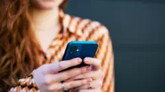 На темном фоне стоит девушка с рыжими волосами в оранжевой рубашке в клетку. В руках девушка держит темно-синий смартфон. На безымянных пальцах девушки по кольцу.