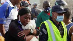 Una mujer está llorando mientras abraza un oso de peluche. A su lado está un policía, con un peto color amarillo fluorescente y tapabocas.