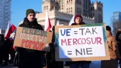 Grupo protesta contra acordo Mercosul-UE em Varsóvia, na Polônia. Homem e mulher seguram cartazes