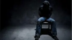 Man wey dem tie im hand behind im back siddon ontop chair