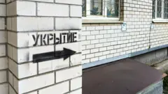 На білій цегляній стіні чорною фарбою нанесено трафаретний напис російською "укриття" та стрілка, що вказує праворуч. Поруч – вікна з ґратами та частина металевого накриття біля фундаменту.