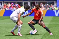 Duelo entre dos jugadores en un partido de fútbol, con un futbolista vestido de rojo que conduce el balón frente a un rival con uniforme blanco, bajo la mirada del público en las gradas.