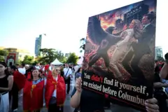 Tiran'da 10 Haziran'daki protesto gösterisinde Arnavutluk bayrağına sarılmış iki kadın ve elinde Aranvutluk sembolü bir kartalın Ivanka Trump ve Jared Kushner'i ittiğini gösteren bir poster: 'Siz bulmadınız, biz Columbus'tan önce de vardık" yazıyor