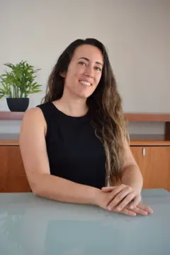 Plano médio da especialista Anna Grifols, vestindo uma camiseta preta, com as mãos sobre uma mesa de vidro e sorrindo para a câmera.