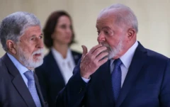 Foto mostra Celso Amorim e Lula, ambos usando ternos escuros e gravatas, conversando. Lula está à direita e coloca a mão sobre a boca para falar a Amorim