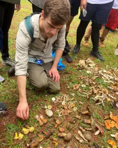 Domnauer arrodillado en el césped examinando una variedad de hongos dispuestos en el suelo, rodeado por otras personas de pie.