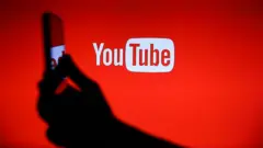 گوشی موبایل به دست مقابل لوگوی یوتیوب با زمینه قرمز