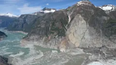 Uma vasta extensão de mar gelado atravessa uma passagem estreita entre montanhas. Grande parte das encostas íngremes das montanhas está desprovida de vegetação verde, revelando rochas cinzentas e marcadas. Os cumes das montanhas estão salpicados de neve e o gelo cintila no fiorde abaixo.