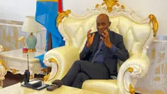 Jean-Jacques Purusi, Guverineri w'intara ya Kivu y'Epfo washyizweho n'ubutegetsi bw'i Kinshasa, mu kiganiro n'ibiro ntaramakuru Reuters i Bukavu, ku itariki ya 12 Gashyantare (2) mu 2025. Yicaye mu ntebe nini y'imisego, yambaye ikositimu y'umukara na karuvati y'ubururu irimo amabara mato y'umweru, akoze ikimenyetso n'ibiganza byombi. Iruhande rwe hari ibendera rya Repubulika ya Demokarasi ya Congo, imbere ku meza hari icyuma gifata amajwi, telefone n'icyombo.