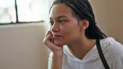 Cathy tiene el cabello oscuro recogido en trenzas y lleva puesto un top blanco, con paredes beige y una ventana de fondo.