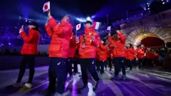 赤い上着に黒っぽいパンツ姿の日本選手らが日本やイタリアの国旗を手に歩いている。後方にはアーチ型のゲートが見える。会場は全体的に暗く、青みがかった照明が照らしている