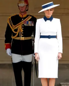 Melania Trump usa um vestido branco com cinto azul e um chapéu branco. Atrás dela está um guarda