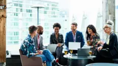 Grupo de pessoas conversando com roupas de trabalho, laptops no colo e copos de cerveja na mesa