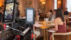 Câmera de filmagem com duas telas filmando três atores sentados junto a uma mesa, em um restaurante sofisticado