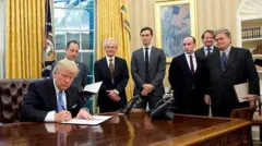 Trump assina documentos na mesa presidencial da Casa Branca, observado por seis assessores de pé