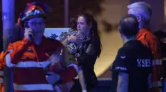 Una mujer toma en brazos a un niño cubierto por una mana isotérmica entre varios miembros de los equipos de emergencia.