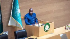 Perezida w'u Burundi Évariste Ndayishimiye arimo kuvuga ijambo mu ndangururamajwi ebyiri, mu cyumba cy'inama cyitwa 'Nelson Mandela Hall', mu nama isanzwe ya 39 y'inteko y'umuryango w'Ubumwe bw'Afurika (AU), ku cyicaro cy'uwo muryango i Addis Ababa muri Ethiopia, ku wa gatandatu, ku itariki ya 14 Gashyantare (2) mu mwaka wa 2026. Yambaye ikoti ry'ubururu ry'amaboko maremare n'ikora rigufi. 