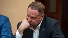 Крупний план: Єрмак в темному піджаку й білій сорочці сидить за столом, спираючи підборіддя на руку. На піджаку — маленький значок із прапором України.