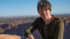 Sentado com uma paisagem ao fundo, o Prof Brian Cox veste uma blusa verde e olha diretamente para a câmera