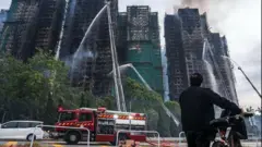 Awọn panapana n na ọpa omi si oke fiofio, sara awọn ọpọ ileesẹ alaja pupọ ti ina alatagba n jo nilu Hongkong, ọkọ ileesẹ panapana kan wa nilẹ, ti ọkunrin kan to wọ asọ kootu dudu si duro, o fa ọkada kan lọwọ, o n wo ile to n jo naa.