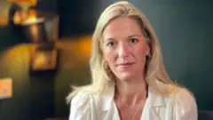Half-length portrait of Fiona Rugg in a white blouse and gold hoop earrings. She has long blonde hair and blue eyes and smiles slightly at the camera. Behind her is a dark green wall with framed pictures and warm lighting.