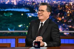 Stephen Colbert sentado y sonriendo en el set, con una taza sobre el escritorio.