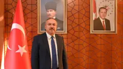 Ali Fidan lacivert bir takım elbise, beyaz gömlek ve mavi bir kravat takmış. Solunda Türk bayrağı duruyor, arkasındaki duvarda Atatürk portresi sağ tarafta da Erdoğan portresi asılı.