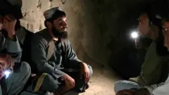 رجل ملتحٍ يرتدي سترة طويلة داكنة وسروالاً تقليديين، ويضع قبعة، يجلس متربعاً على أرضية عارية داخل كهف مظلم، يظهر في يسار الصورة وهو ينظر إلى رجل يجلس إلى اليمين يحمل مصباحاً يدوياً.