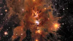 Nuvens alaranjadas rodopiantes são vistas contra um pano de fundo de estrelas distantes, com três centros brilhantes de formação estelar brilhando em uma linha vertical.