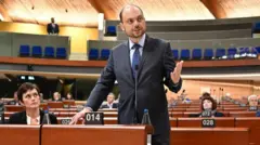 Худой мужчина в костюме выступает в парламенте. На него смотрит его жена, сидящая рядом