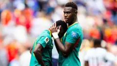 Lamine Camara, du Sénégal, appuie son visage contre la poitrine de son coéquipier Moussa Niakhate, réconforté après l'élimination face à la Belgique en seizièmes de finale de la Coupe du Monde 2026 à Seattle. Niakhate a les mains jointes derrière la tête de Camara pour le serrer contre lui. Tous deux sont visibles jusqu'à la taille et portent leurs maillots verts du Sénégal, ornés de liserés jaunes et rouges. L'arrière-plan est très flou, laissant apparaître un joueur belge de dos et la foule.
