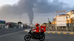 عامل توصيل على دراجة نارية حمراء، فيما يتصاعد خلفه عمود كثيف من الدخان عقب انفجار في المنطقة الصناعية بالفجيرة في 3 مارس/آذار 2026.