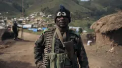 Umusirikare wa FARDC yifotoje ari mu kigo cya gisirikare cy'i Lusogha, hafi y'umurongo w'imbere wo ku rugamba, muri kilometero zirenga 10 mu majyepfo ya Kanyabayonga, mu ntara ya Kivu ya Ruguru, mu burasirazuba bwa Repubulika ya Demokarasi ya Congo. Ifoto yo ku itariki ya 14 Gicurasi (5) mu mwaka wa 2024. Arahagaze, yambaye ingofero y'ubudodo n'imyenda ya gisirikare, afite imbunda n'umuzingo w'amasasu ku rutugu. 
