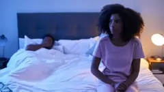 A woman sits anxiously on the edge of her bed as a man lies in bed rubbing his face