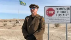 Un hombre de bigote, uniformado con un abrigo y boina color marrón y gafas oscuras, está para parado con pose oficial frente a un aviso que lee: "Alto. Se requiere permiso para entrar". En el fondo ondea una bandera verde y el paisaje es desértico