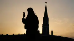 A person is silhouetted against a glowing orange sky as they look at their smartphone, with the silhouette of the Kremlin in the background during sunset in the Red Square in Moscow on 31 March.