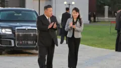 Kim Jong Un, de terno escuro, camisa branca e gravata vermelha, e Kim Ju Ae, com os longos cabelos presos, usando uma jaqueta cinza curta, camisa e calça pretas, aplaudem enquanto se afastam de um grande carro preto com a placa 7. 27.1953, data da assinatura do Acordo de Armistício Coreano.
