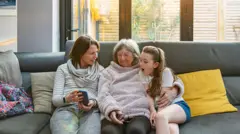 Três gerações de mulheres de uma mesma família sentadas no sofá, observando uma foto no celular