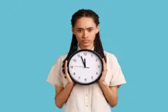 Femme avec des dreadlocks tenant une grande horloge murale, regardant l’appareil photo avec des émotions désagréables, il est temps d’y aller 