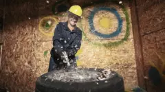 Kobieta rozbija szklany pojemnik w pokoju gniewu. Jest ubrana w odzież ochronną i kask chroniący przed rozbijaniem i niszczeniem przedmiotów. Wnętrze siłowni Rage Room w centrum Hamilton w Ontario w Kanadzie.