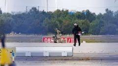 黒い長袖長ズボン姿の人物が、空港の駐機場を歩いている。右隣にベージュのロボットが見える