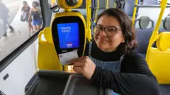 Mulher passa seu bilhete único na catraca do ônibus em São Paulo e a catraca exibe na tela a mensagem "Tarifa zero". Ela tem cabelos escuros curtos, usa óculos redondos, veste camiseta preta de mangas compridas e um macacão jeans, e está sorrindo discretamente para a câmera. 