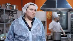 Yulia lleva un mono de trabajo y una redecilla para el pelo en una cocina industrial con paredes naranjas. Un hombre con gorro y delantal de chef camina detrás de ella.