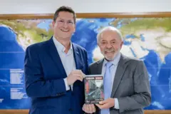 Jilmar Tatto presenteando o presidente Lula com seu livro "Tarifa zero: Embarque nesta ideia". Ambos estão segurando o livro e sorrindo, em frente a um mapa-múndi