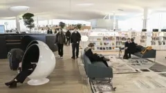 Interior de uma biblioteca moderna, com iluminação natural e grandes janelas. Há estantes baixas cheias de livros organizados, sofás e mesas onde pessoas leem, conversam ou usam dispositivos eletrônicos. À esquerda, uma pessoa está sentada dentro de uma cadeira em formato de cápsula branca, parcialmente fechada. No centro, duas pessoas caminham pelo espaço. O ambiente tem design contemporâneo, com piso de madeira, tapetes e plantas distribuídas pelo local.